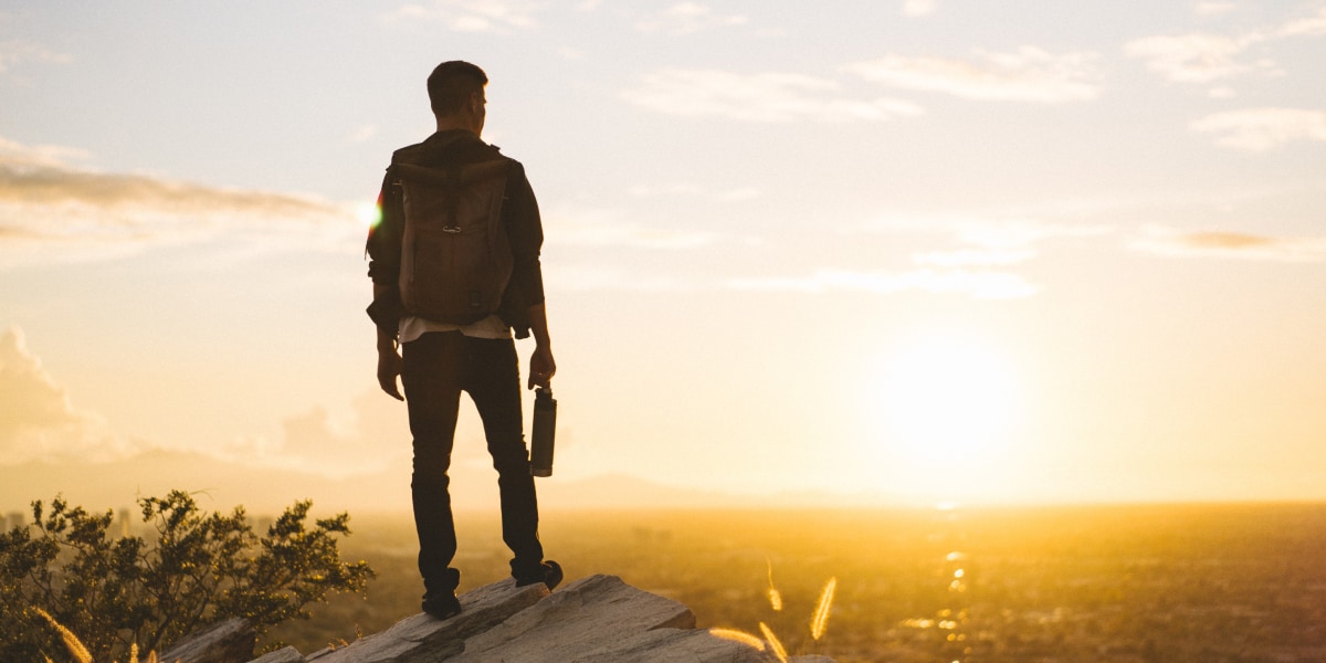 Ein Mann auf einer Klippe stehend, wie er auf das darunter liegende Tal und den Sonnenuntergang schaut