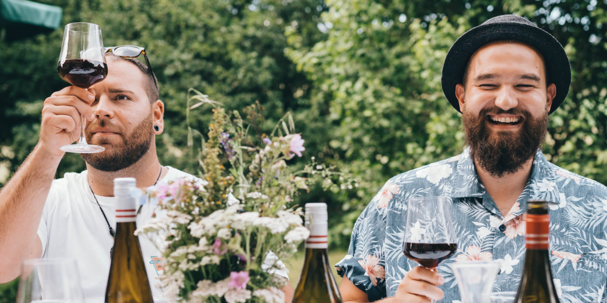 Zwei Männer lachend in einem Garten bei einem Glas Wein