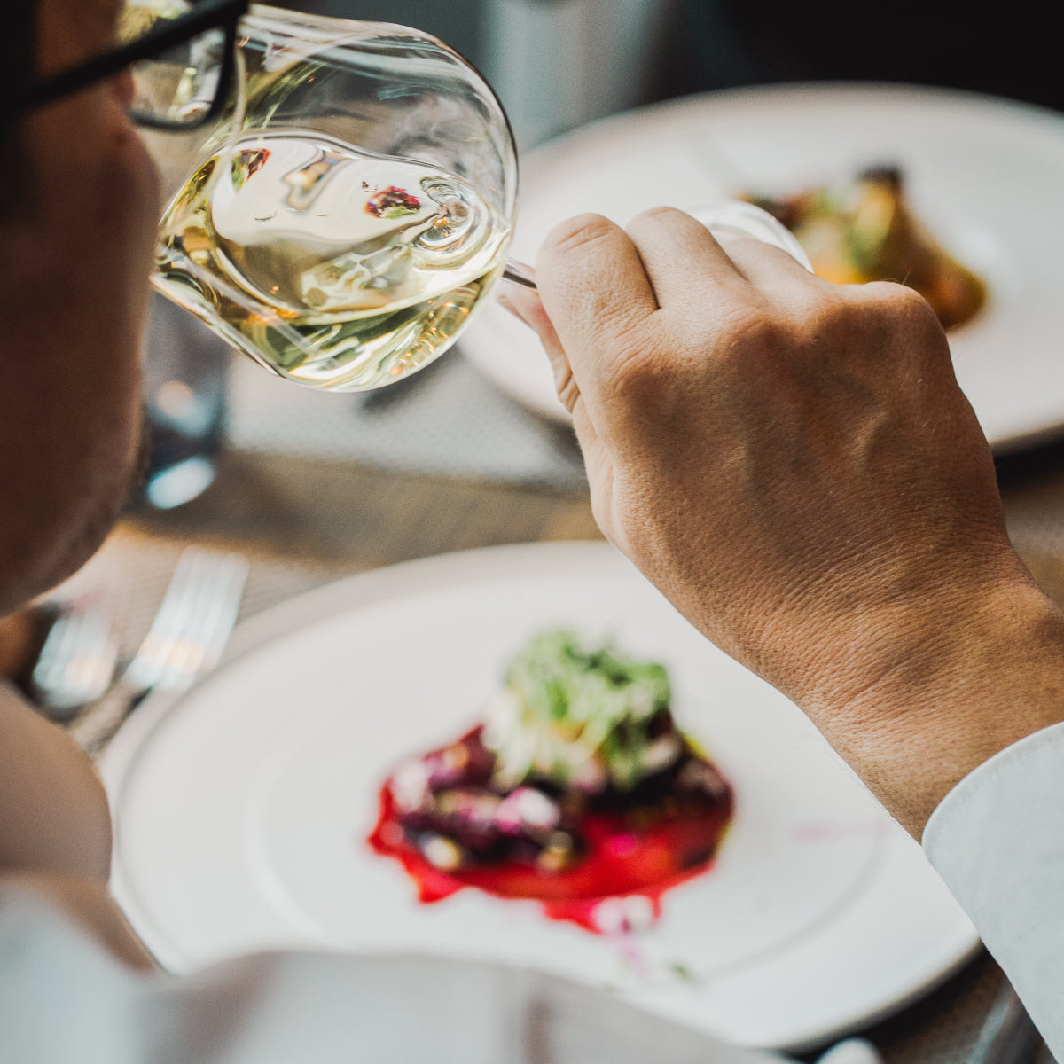Ein Mann an einem gedeckten Tisch sitzend, wie er ein Glas Weißwein trinkt und vor sich einen Teller mit Essen stehen hat
